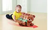 A baby is crawling on the floor, reaching for a toy bear that is also crawling. The bear has motion blur to indicate movement. Produktbild-Vorschau