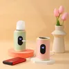 Two portable bottle warmers, one with a baby bottle, are displayed on a light orange and white surface, with a vase of pink tulips in the background. A red power bank is in the foreground. Produktbild-Vorschau