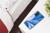 A smartphone with a blue abstract design case lying on a white surface next to a plant and a bed. Produktbild-Vorschau