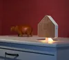 A wooden and white house-shaped sound machine with a light at the base sits on a white dresser next to a wooden hippo figurine. The background is a solid red wall. Produktbild-Vorschau