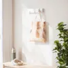 A tote bag with a stone-like pattern hangs on a white folding hook mounted on a textured wall. A water bottle and headphones sit on a wooden bench below. Podgląd zdjęcia produktu