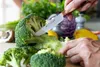A person is cutting broccoli with a knife in a kitchen setting with other vegetables in the background. Voir les images du produit