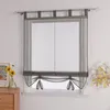 A sheer gray Roman blind is installed on a window, with decorative ties at the bottom. A small vase with yellow flowers sits on a wooden dresser to the left, and a branch with yellow leaves is on the right. Produktbild-Vorschau