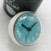 A close-up shot of a small blue clock with black hands, placed on a white surface next to a dark pot with text. Produktbild-Vorschau