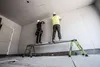 Two construction workers are installing drywall on a wall, standing on a telescopic plank. Produktbild-Vorschau