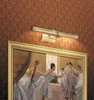 A picture light illuminates a painting of ballerinas on a patterned wallpapered wall. Podgląd zdjęcia produktu