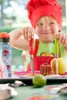 A child wearing a chef's hat and apron holds two knives, with food and a water bottle on a table. Produktbild-Vorschau