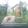 A hand holds a wooden house-shaped frame with a plant icon inside, against a blurred green background. Produktbild-Vorschau