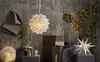 A festive display of Christmas decorations, including illuminated paper stars, candles, and wreaths, set against a textured concrete wall. Produktbild-Vorschau