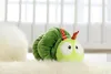 A green plush caterpillar toy lies on a fluffy white rug in front of a blurred background of white blinds. Produktbild-Vorschau