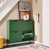 A dark green shoe cabinet with gold legs is placed in a hallway next to a staircase. An open compartment reveals shoes inside. A piece of abstract art and a white vase are on top of the cabinet. A yellow stool is to the left, and a rug is on the floor. Náhľad obrázku produktu