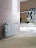 A white DeLonghi stand heater is mounted on a light-colored wall in a living room with a plush carpet and a bookshelf in the background. Produktbild-Vorschau