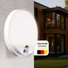 A split image showing a white LED outdoor wall light with a motion sensor on the left, and a modern wooden house with large windows on the right. Produktbild-Vorschau