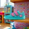 A child's turquoise shoe with pink accents is placed on a wooden shelf in a playroom setting. The background features colorful wallpaper with rainbows and toys. Produktbild-Vorschau