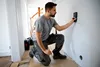 A man is kneeling on the floor in a hallway, using a stud finder on a white wall. Voir les images du produit