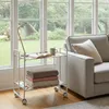 A white rolling cart with two shelves is placed next to a sofa in a room with large windows. Voir les images du produit