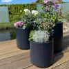 Three dark gray planters with various flowers and greenery are arranged on a wooden deck next to a body of water. Podgląd zdjęcia produktu