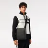 A man wearing a black and white jacket and black pants stands against a white background, holding a black messenger bag. Produktbild-Vorschau