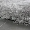 A close-up, angled view of a shaggy, gray rug with a high pile, showing the texture of the fibers and the edge of the rug resting on a tiled floor. Produktbild-Vorschau