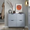 A narrow shoe cabinet with four flip-down drawers is displayed in a hallway with a coat hanging on a hook to the left and artwork on the wall above the cabinet. Náhľad obrázku produktu