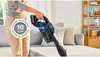 A person is using a cordless vacuum cleaner in a living room setting, with a sofa and shelving unit visible. Náhled obrázku produktu