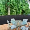 A patio table and chairs are set up on a wooden deck, with a dark privacy fence behind them and trees in the background. Produktbild-Vorschau