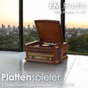 A vintage-style record player and radio sits on a polished floor in front of a large window overlooking a city skyline at sunset. Produktbild-Vorschau