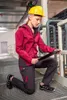 A woman in workwear is kneeling on a metal grate walkway, looking at a clipboard. Produktbild-Vorschau