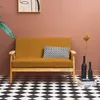 A mustard yellow sofa with two decorative pillows sits on a black and white checkered floor in front of a terracotta-colored wall. A plant is visible on the left, and a dried flower in a vase with two black bottles is on the right. Anteprima dell’immagine prodotto