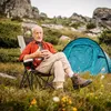 An older man sits in a camping chair next to a tent in a mountainous, grassy area. Produktbild-Vorschau