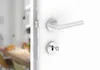 A close-up view of a silver door handle and lock mechanism on a white door, with a blurred interior scene visible through the open doorway. Produktbild-Vorschau