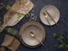 A rustic table setting with plates, cutlery, a napkin, a wooden cutting board with beans, and greenery. Produktbild-Vorschau
