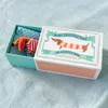 A small stuffed dachshund dog wearing a striped sweater, lying in a teal box with a polka dot lining. The box is partially open, revealing the product inside. The lid of the box is decorated with a cartoon illustration of a dachshund and text. Produktbild-Vorschau