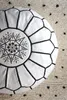 A close-up view of a white leather pouf with black stitching and embroidery, placed on a textured rug with black speckles. Produktbild-Vorschau