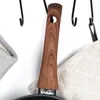 A close-up of a frying pan handle with a wooden texture, hanging on a hook. Two fabric pot holders are visible on either side. Podgląd zdjęcia produktu
