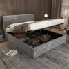 A gray upholstered bed with its storage compartment open, showing pillows and folded blankets inside. The bed has a diamond-stitched headboard and is placed on a gray rug in a room with wooden walls and curtains. Anteprima dell’immagine prodotto