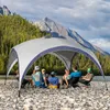 A group of people are sitting under a canopy tent on a rocky riverbank, with a forest and mountains in the background. Anteprima dell’immagine prodotto