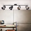 A black ceiling spotlight fixture with four adjustable lamps is mounted on a white ceiling above a wall with a textured gray finish. Below the fixture, a wooden shelf holds a globe and a stack of books. Produktbild-Vorschau