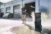 A man is pressure washing the ground in front of a building with a car parked nearby. A stack of toolboxes is to his right. Náhled obrázku produktu