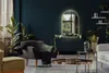 A modern living room with a dark teal wall, featuring a large arched mirror with LED lighting, a dark green dresser, a grey armchair with a black throw, and a coffee table with a copper-toned top. Produktbild-Vorschau