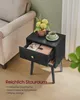 A black nightstand with an open drawer, a cup of coffee, and a vase of flowers next to a sofa. Produktbild-Vorschau