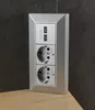 A silver electrical outlet with two USB ports and two standard sockets, mounted on a dark gray textured wall above a light wood surface. Anteprima dell’immagine prodotto