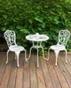 A white metal bistro set with two chairs and a table is placed on a wooden deck, with a lush green hedge in the background. Náhled obrázku produktu
