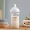 A glass baby bottle filled with milk, placed on a wooden surface with baby toys and folded blankets in the background. Podgląd zdjęcia produktu