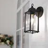 A black outdoor wall lantern with a clear glass enclosure is mounted on a white paneled wall next to a window. A bouquet of flowers is visible in the lower left corner. Voir les images du produit