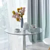 A round glass-top dining table with a silver metal base, set with two cups of coffee, a teapot, and a vase of flowers, placed near a window with sheer curtains and a green gradient curtain. Produktbild-Vorschau