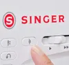 A close-up shot of a Singer sewing machine with a finger pressing a button. Produktbild-Vorschau