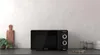 A black microwave oven is placed on a countertop in front of a textured wall. There are some kitchen accessories to the right and a plant to the left. Anteprima dell’immagine prodotto