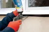 A person wearing orange gloves is applying expanding foam from a can to seal a gap around a window frame. Produktbild-Vorschau
