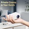 A hand holding a white mini projector in a living room setting. Produktbild-Vorschau